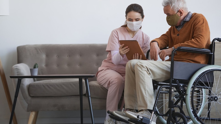 A female health worker shows documents to an older man who is in a wheelchair.