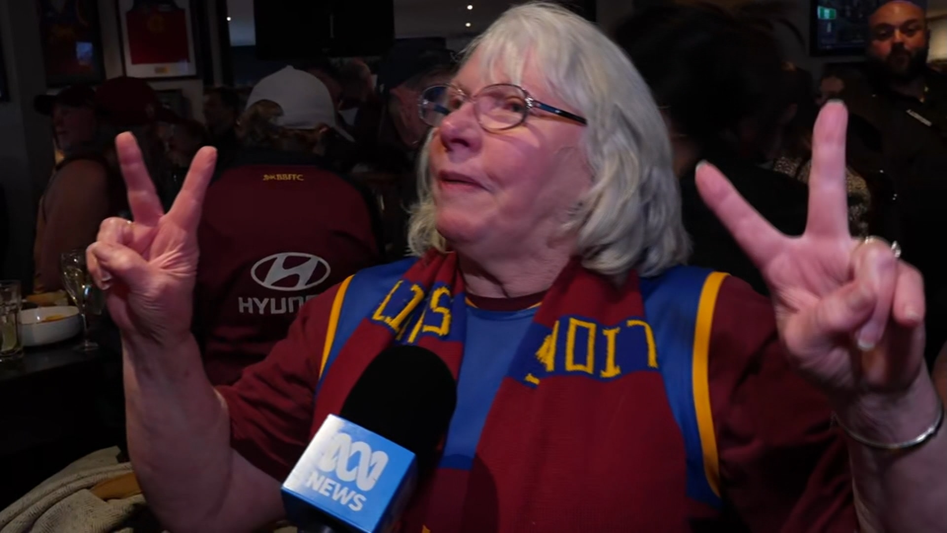 A white haired woman wearing glasses and a Brisbane Lions scarf holds her two hands up with two fingers up.