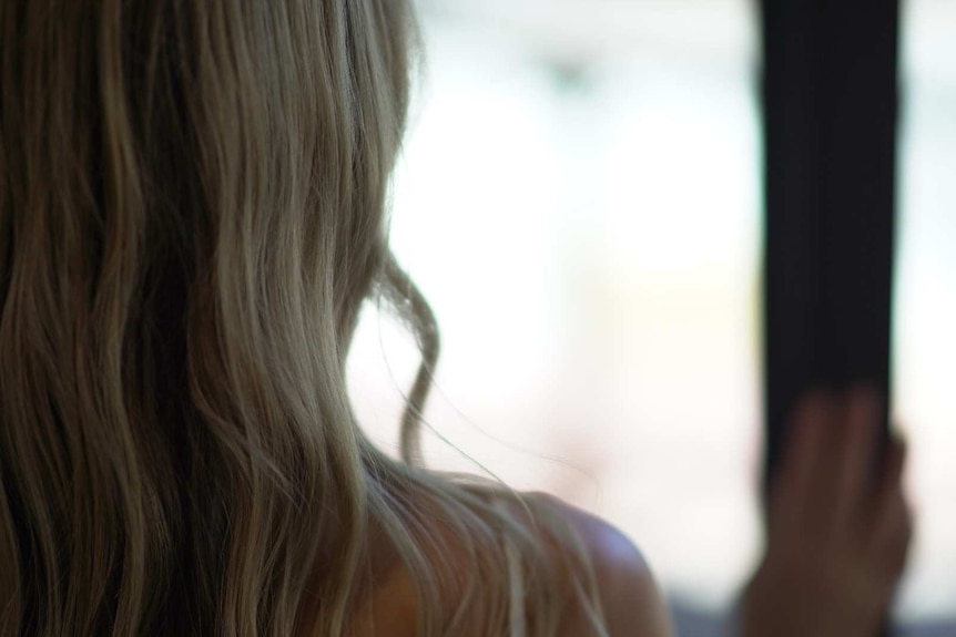 A young woman with long blonde hair is shown from behind looking out a window.