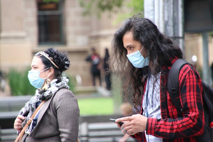 Two pedestrians wait at a set of lights, both wearing face masks.
