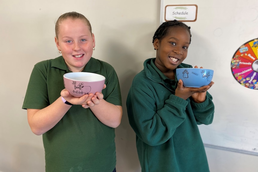 Two smiling girls wearing green school uniforms hold bowls up to the camera