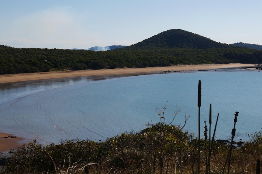 A beach coast line, blue waters, vegetation and a hill and sky in the distance at dusk.