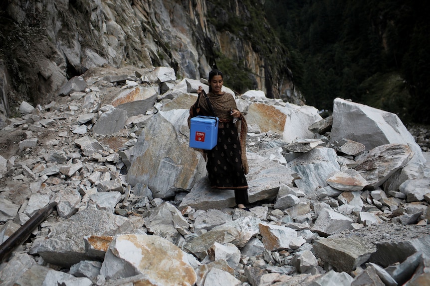 A woman holding a blue container walks over rocky terrain. 