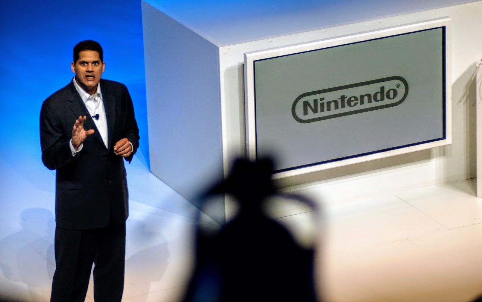 A man stands in a suit giving a presentation with a large Nintendo sign in the background