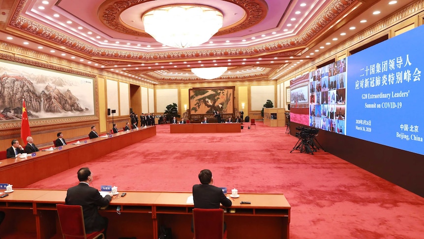 Men sit spread far apart behind a long desk in large room in front of a very big screen.