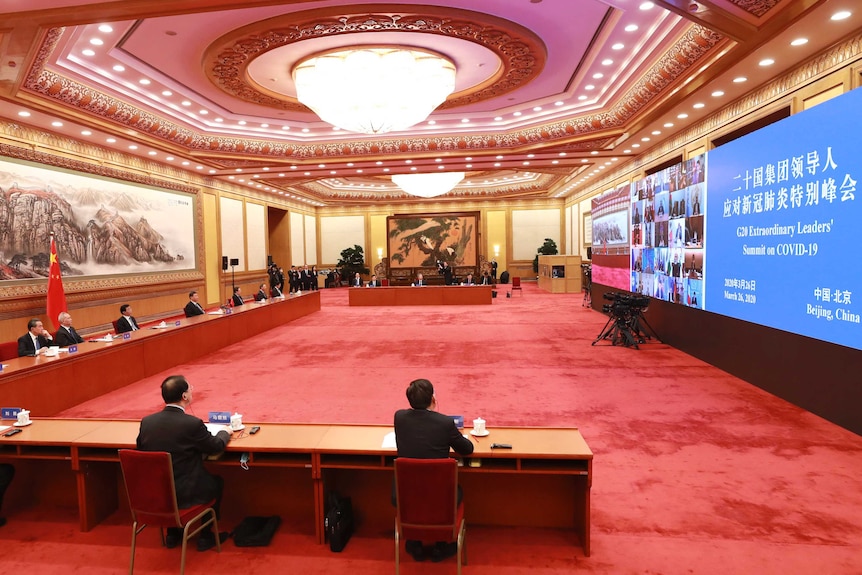 Men sit spread far apart behind a long desk in large room in front of a very big screen.