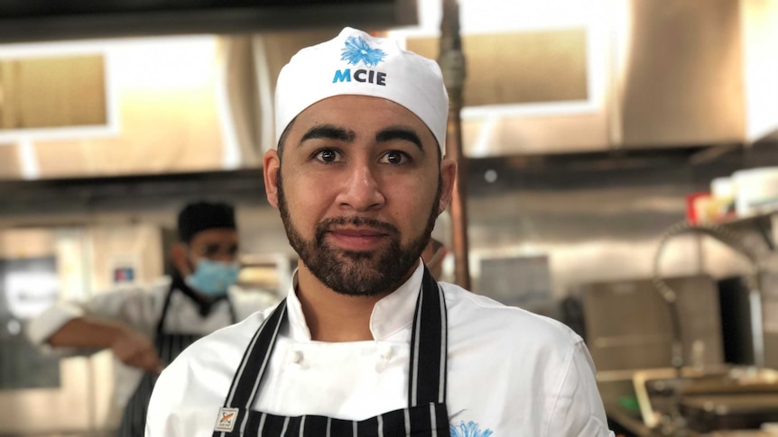 Alejandro Montecimos looks at the camera, while standing in chefs clothes in a commercial kitchen.
