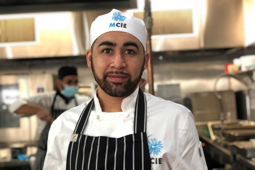 Alejandro Montecimos looks at the camera, while standing in chefs clothes in a commercial kitchen.