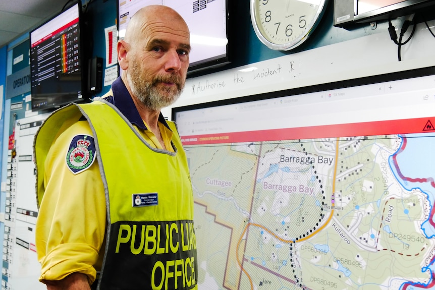 A middle-aged man in high-vis stands in front of a map hanging on an office wall.