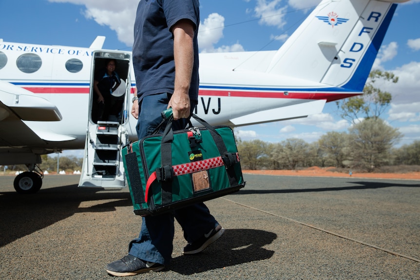 A clinician holding a medical bag stands on the tarmac in front of a Royal Flying Doctor airplane.