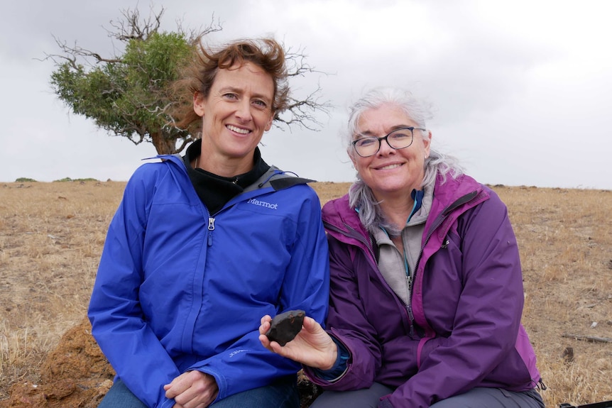 Two women outside looking windswept hold a small chunk of rock.