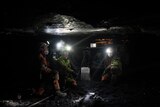 In a pitch black coal mine with a low rocky ceiling, four coal miners wearing bright headlamps sit around machinery.