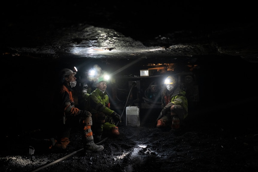 In a pitch black coal mine with a low rocky ceiling, four coal miners wearing bright headlamps sit around machinery.