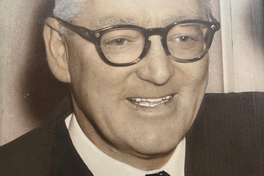 A dated sepia photo of a smiling man with black-rimmed glasses, a suit and tie.