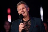 Adam Hills speaks into a microphone onstage at the ParalympicsGB Homecoming