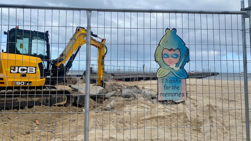 A digger is seen in front of Altona Pier in Melbourne's south-west with a sign on a fence reading 'Thanks for the memories'