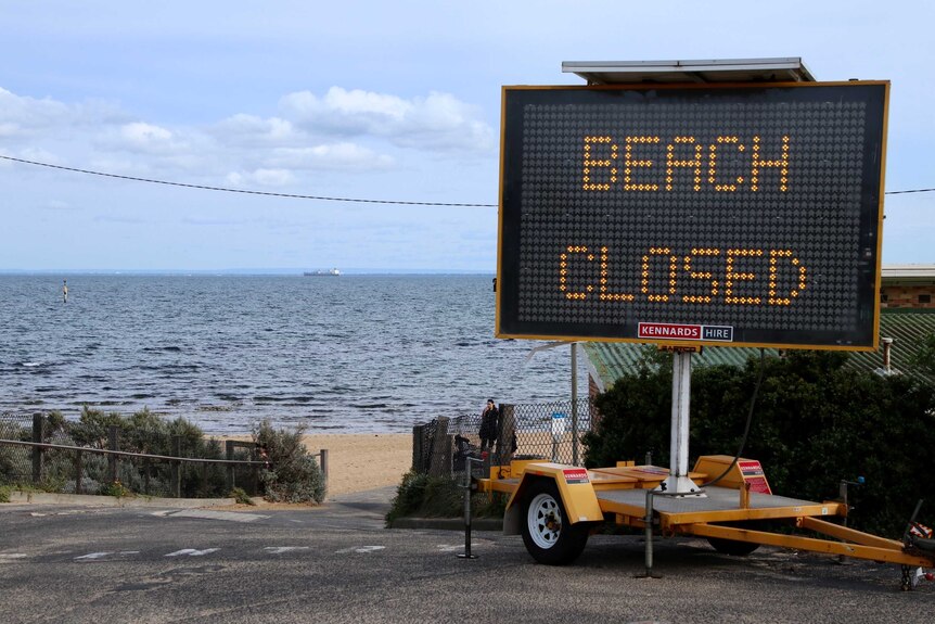 A large electric sign on concrete in front of a beach with a woman on the sand says beach closed.