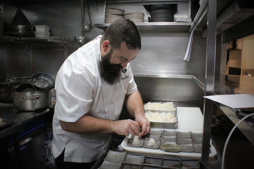 A chef in a white jacket plates up canapés in a stainless steel commercial kitchen.