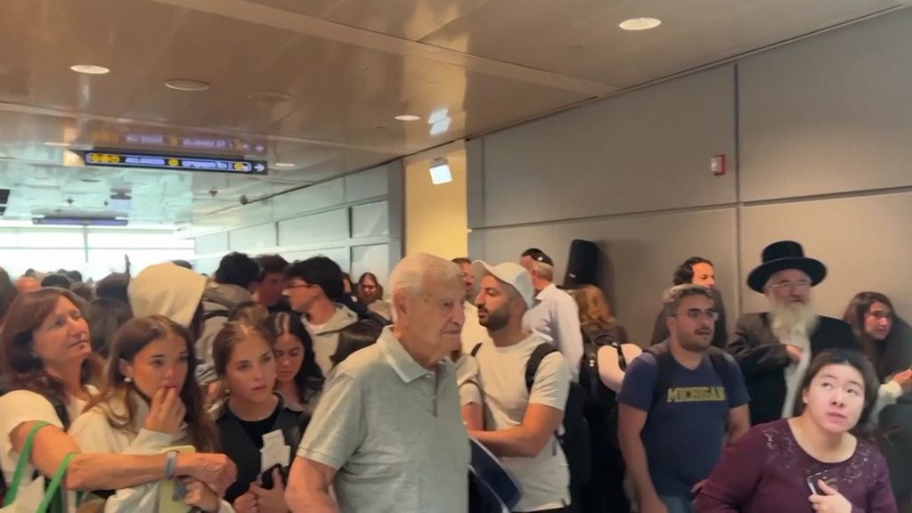 Dense crowd of passengers moving through Tel Aviv airport, concerned looks.