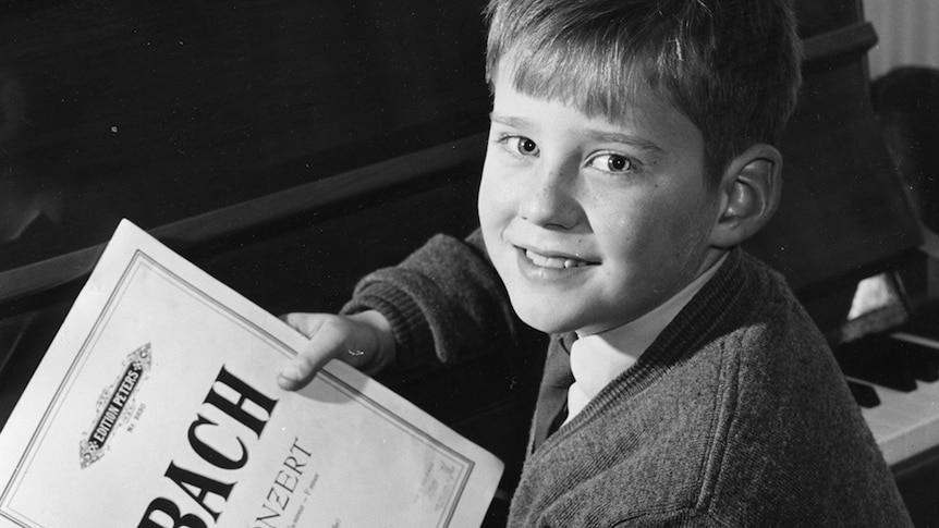 A black and white photo of a young Geoffrey Tozer sitting at a piano, smiling at the camera and holding sheet music by JS Bach.