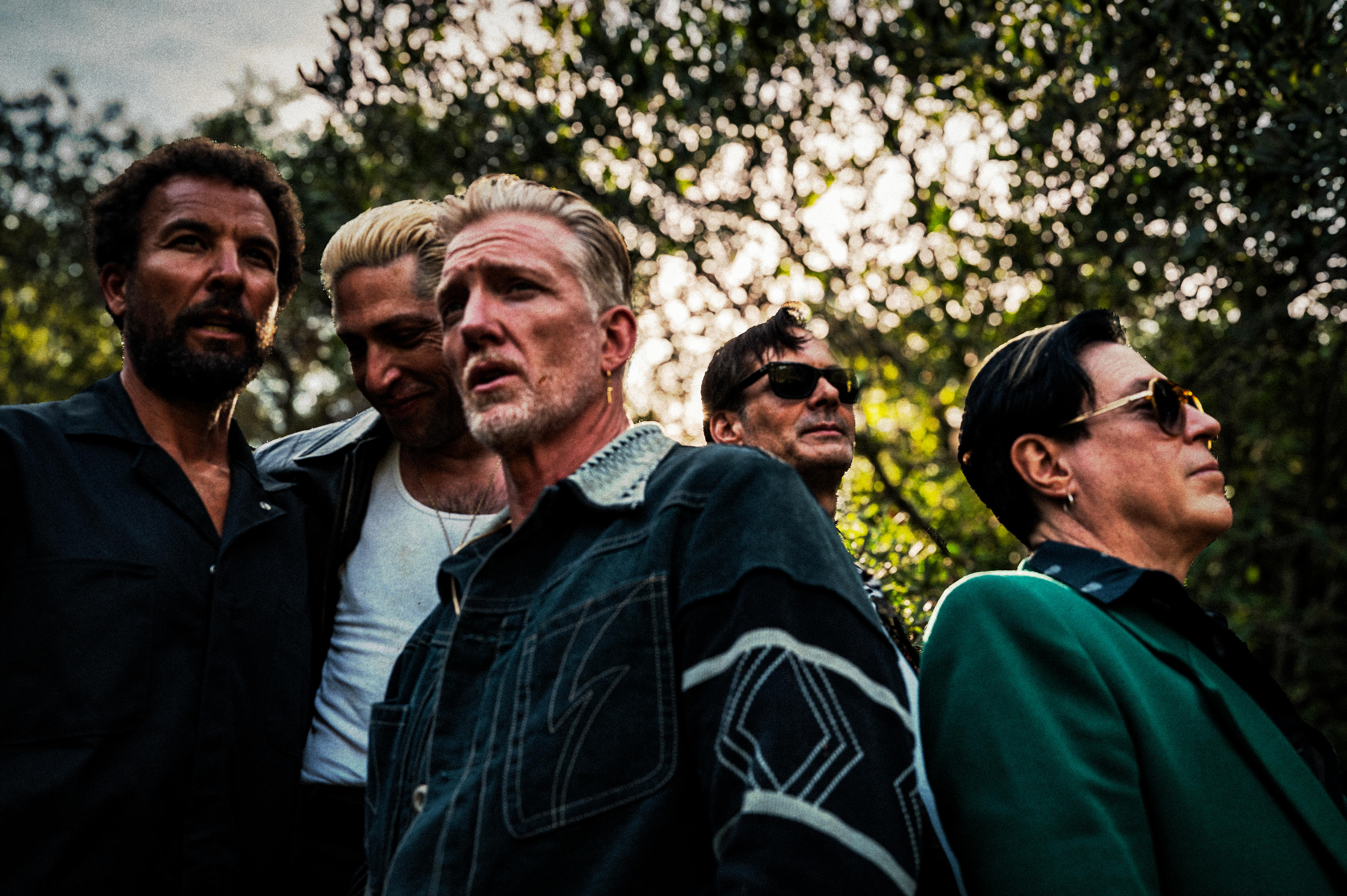 Five members of QOTSA pose outdoors beneath trees, photographed from a low angle with sunlight filtering through leaves