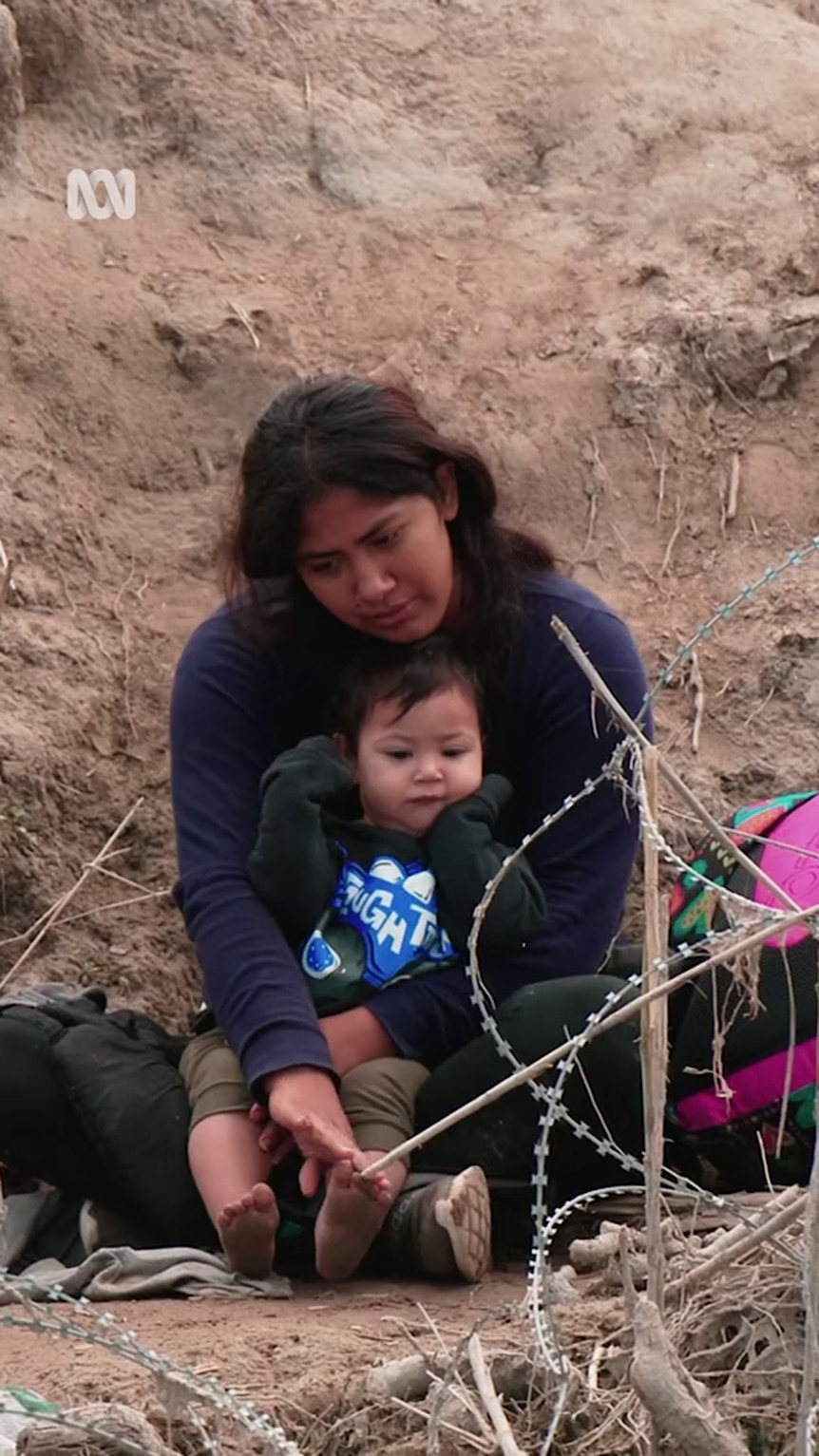 A young brown-skinned woman holds a toddler in her arms as they sit on dirt behind razor wire