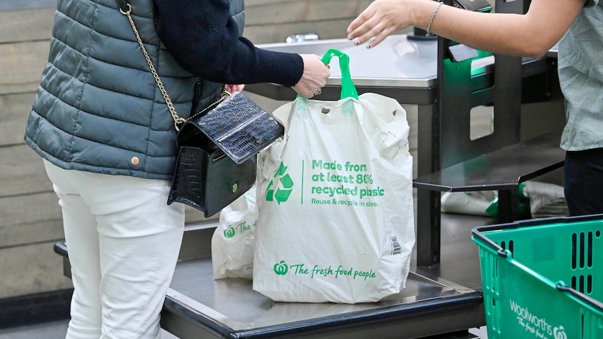 A person grabs a full Woolworths-branded reusable shopping bag from a supermarket check out cashier. 