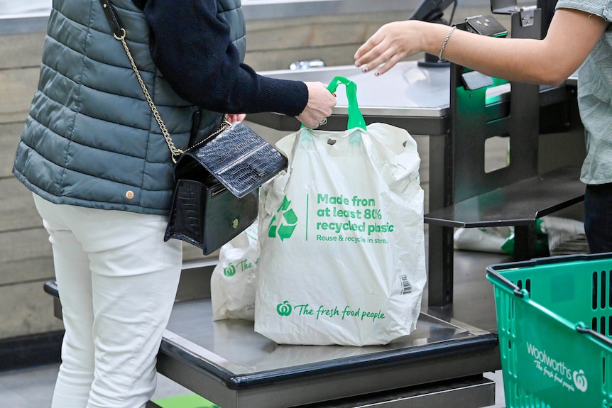 A person grabs a full Woolworths-branded reusable shopping bag from a supermarket check out cashier. 