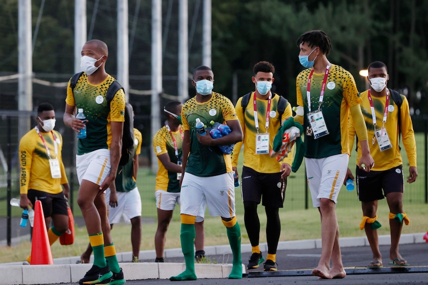 A group of men in South African soccer uniforms and face masks walk down a street near a pitch 