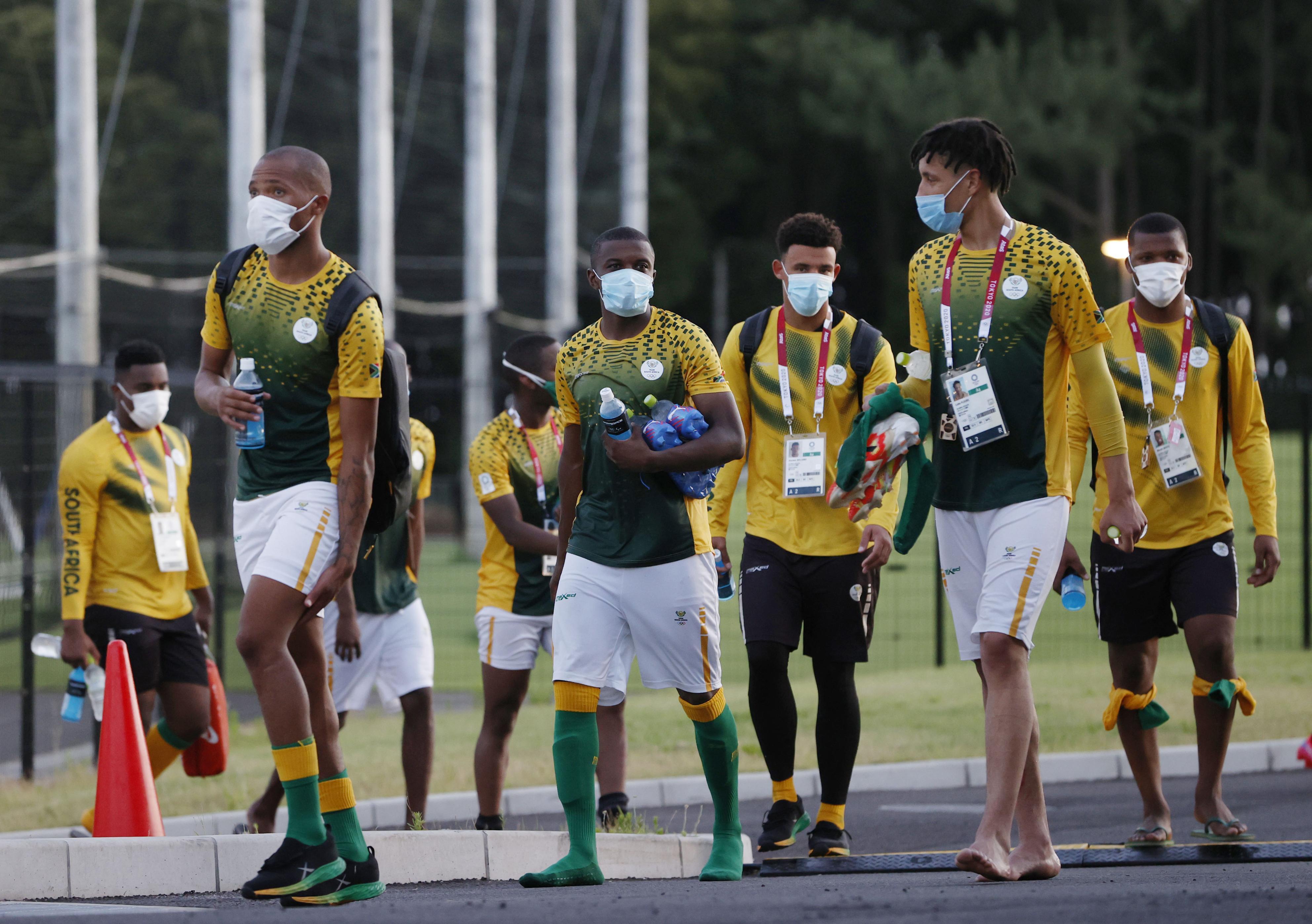 A group of men in South African soccer uniforms and face masks walk down a street near a pitch 