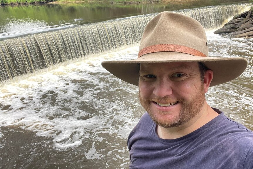a man wearing an akubra hat standing in front of a water dam