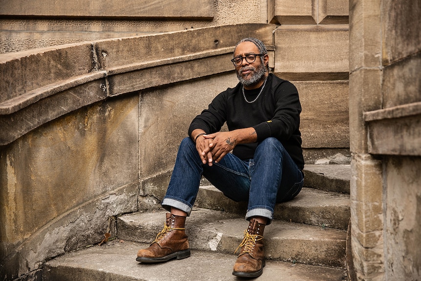 Colour photo of artist Arthur Jafa sitting on stairs in front of sandstone building at the National Art School in Sydney.