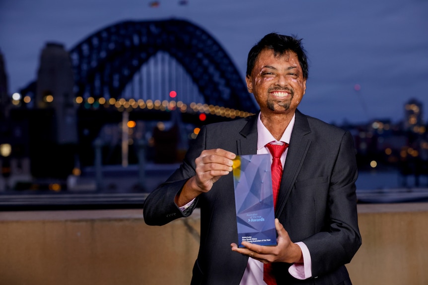 A man in a suit holds up a glass award in front of the Sydney Harbor Bridge.