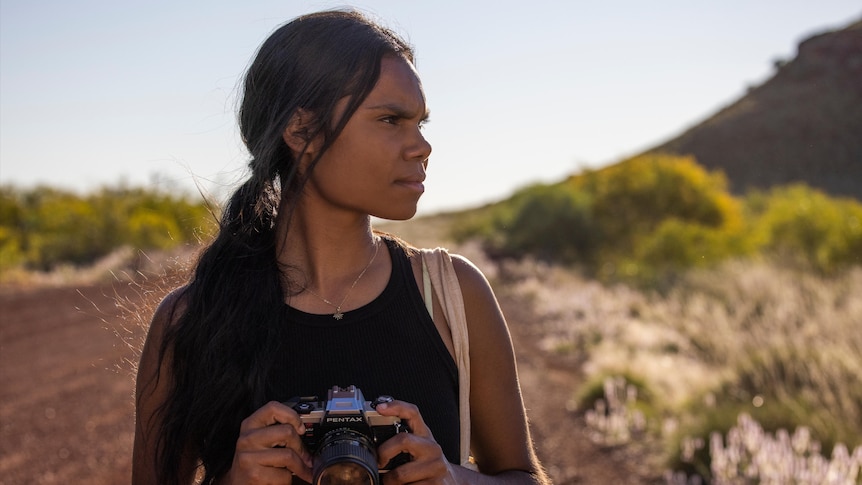 A young Aboriginal woman stands with an analogue camera in her hands