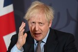 Boris Johnson raises hand and looks forward, hair dishevelled in front of a British flag.