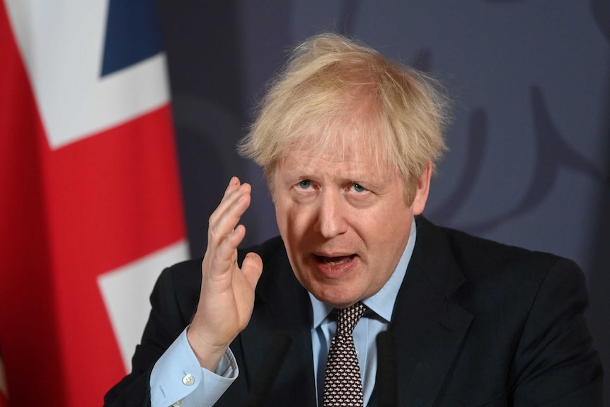 Boris Johnson raises hand and looks forward, hair dishevelled in front of a British flag.