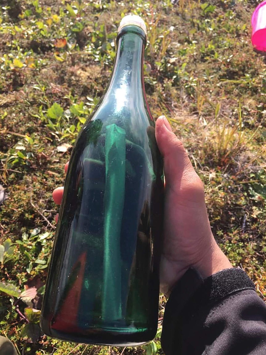 A hand is seen holding a green bottle over grass. The bottle contains a letter.