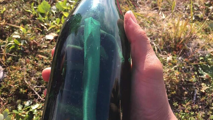 A hand is seen holding a green bottle over grass. The bottle contains a letter.