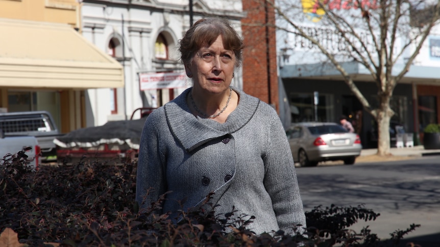 An older woman standing on the main street of a country town wearing a grey coat.