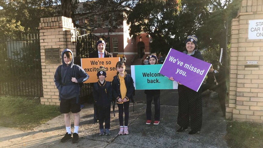 Students stand outside the school gates where teachers hold colourful signs that say welcome back, and we've missed you.