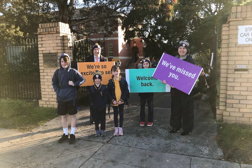 Students stand outside the school gates where teachers hold colourful signs that say welcome back, and we've missed you.