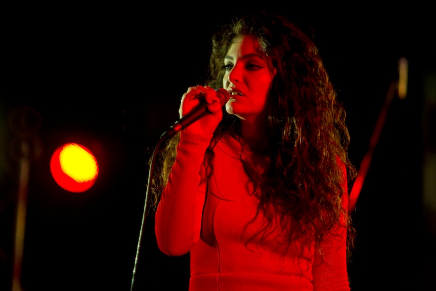 Lorde, bathed in red light, sings into a microphone on stage