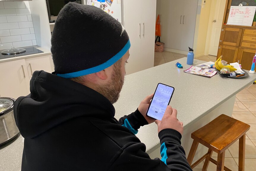 A man wears a black and teal beanie and jumper, he looks at a mobile phone with his electricity bill, he sits at kitchen bench