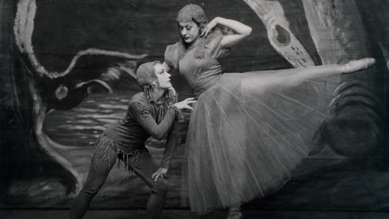 B&W photo of a crouching dancer looking up at another dancer standing with one foot en pointe and the other raised.