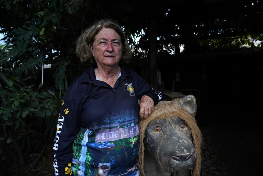 Woman with short, curly hair wearing long-sleeved, dark blue shirt, leans on lion statue with serious expression.