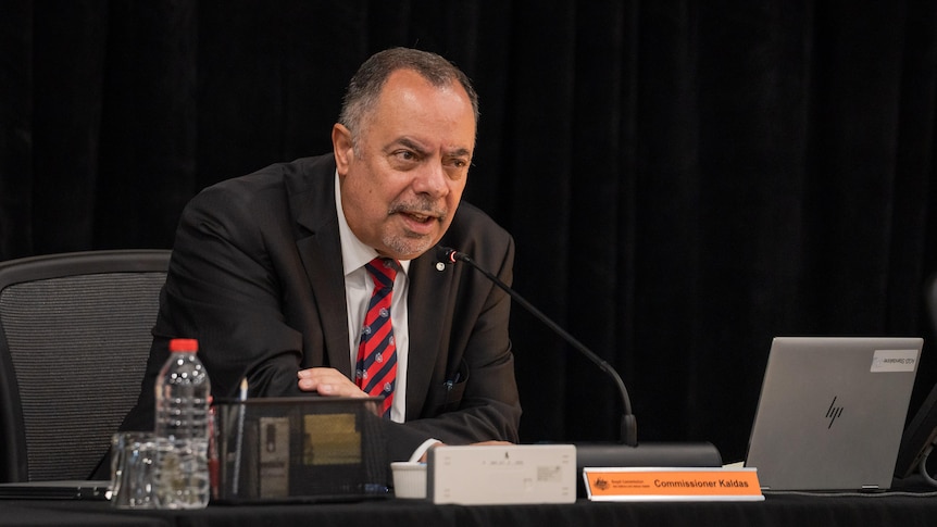 A man in a black suit leans into a microphone on a desk. 