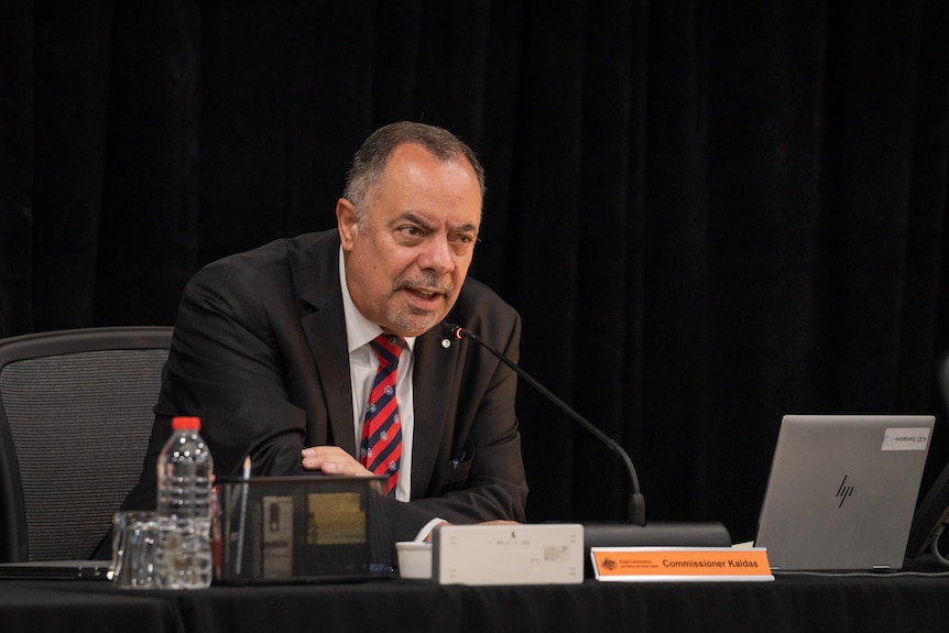 A man in a black suit leans into a microphone on a desk. 