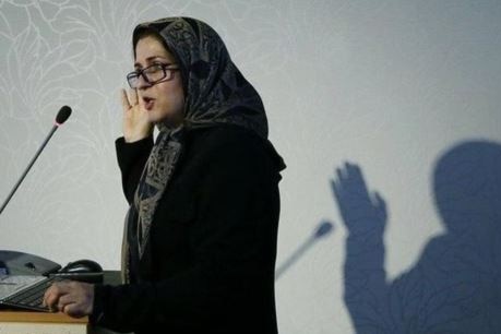 Meimanat Hosseini Chavoshi giving a lecture. She is standing at a podium with a microphone, wearing glasses and a headscarf.