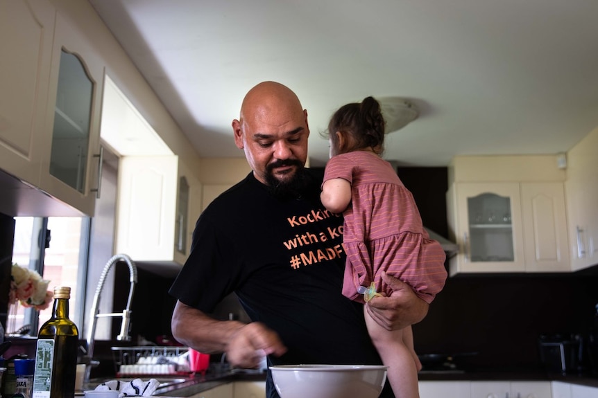 A man holds his daughter at a breakfast bench while he prepares food.