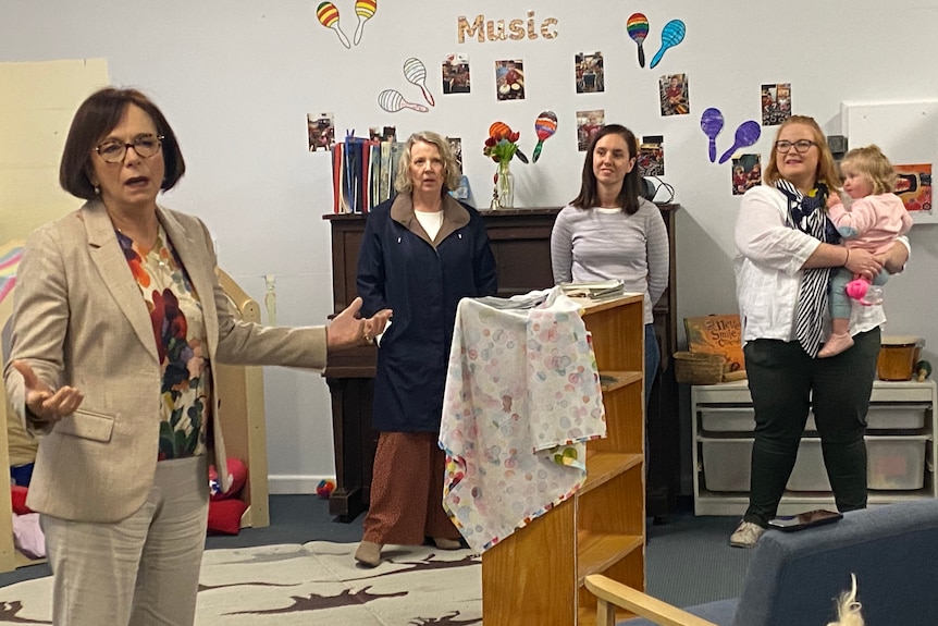 A woman in a cream blazer with dark red hair speaks to a crowd of young mothers, the mother on the right is holding a toddler